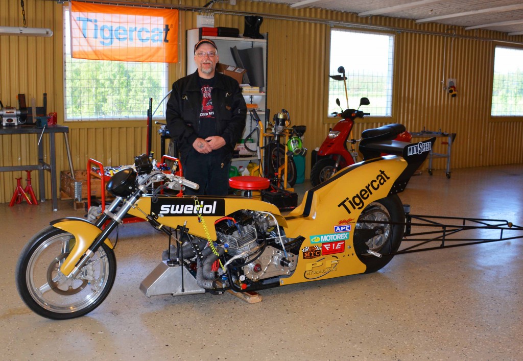 Mats standing with his drag bike inside a workshop, a Mackolines Machines & Hire flag hangs in the window behind him.