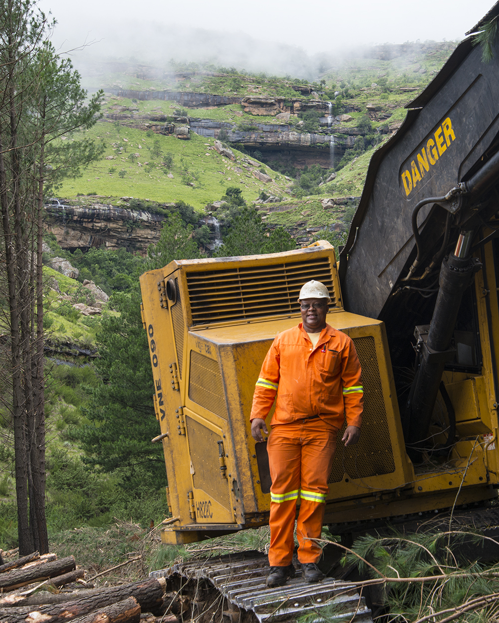 A Mackolines Machines & Hire operator stands proud and smiling on the tracks of his machine in front beautiful hilly green landscape. 