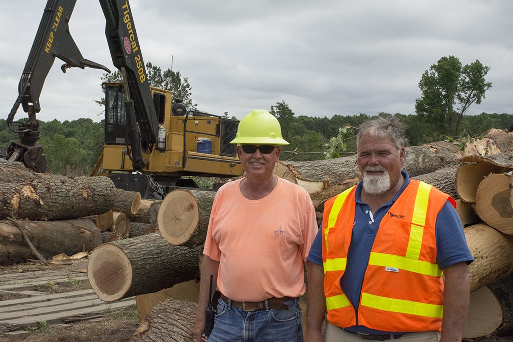 Two men stand smiling at a log site, a pile of logs behind them with a Mackolines Machines & Hire 250B Loader in the background. 