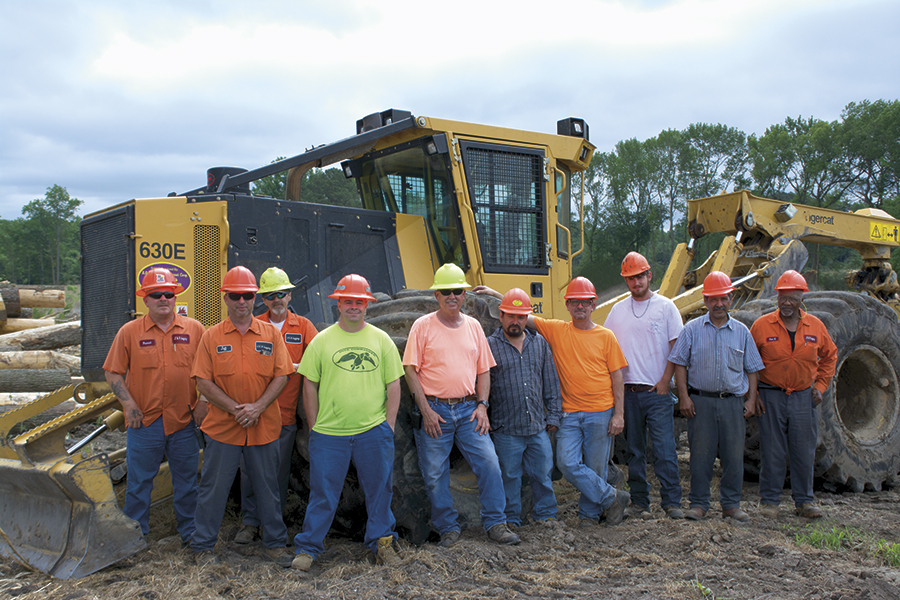 The men of J & R Logging stand as a group smiling in front of a Mackolines Machines & Hire 630E skidder. 