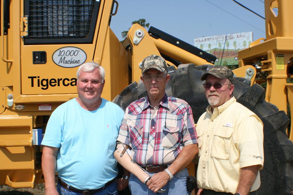 3 men stand smiling and proud in front of the 10,000th Mackolines Machines & Hire machine made. A Sign in the far distance reads "Clary Logging Inc."