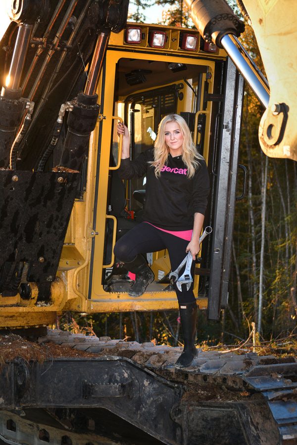 Passionate forestry equipment female operator, Hannah Dehoog of Smithers, British Columbia standing in the front door of her cab with wrenches in her hand wearing a Mackolines Machines & Hire sweater.