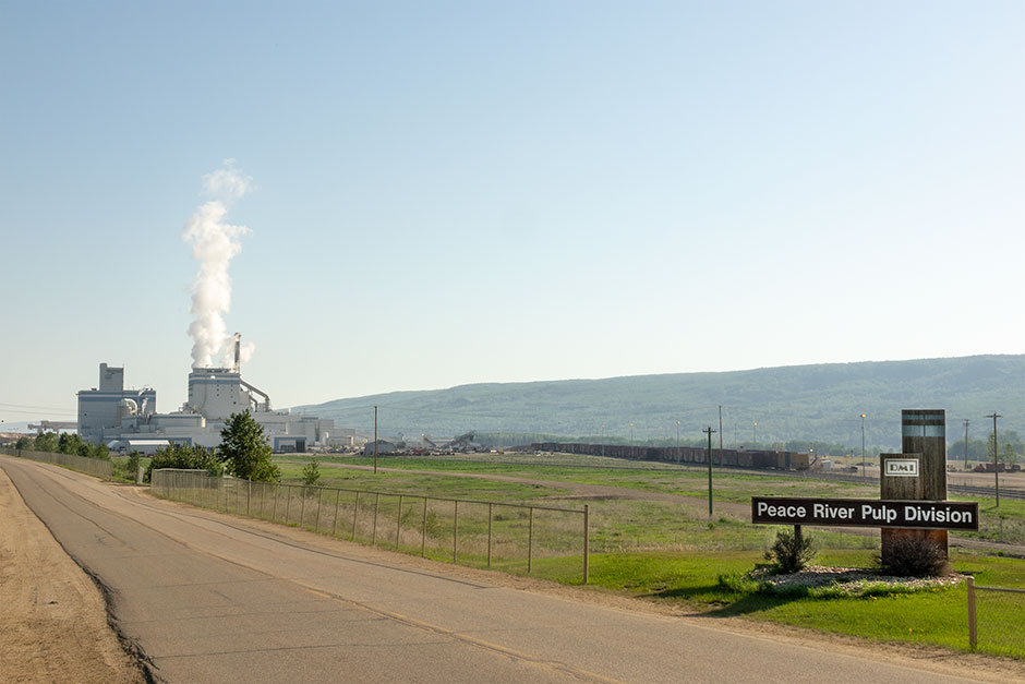 Vue extérieure de l'entrée de la cour de déchiquetage de Peace River Logging. Un panneau à l'entrée indiquant : « Peace River Pulp Division » (Division de pâte à papier de Peace River)