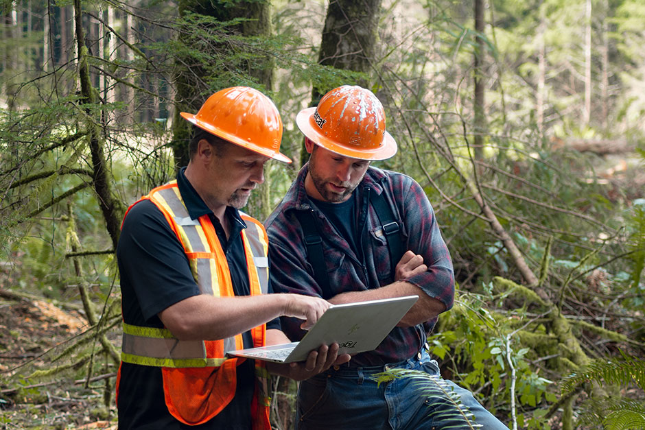 Adam conferring with a Mackolines Machines & Hire representative and looking at a laptop in the forest.